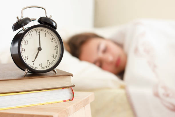 menina de adolescente dormindo em uma cama branca. relógio despertador em primeiro plano em uma pilha de livros - alarm clock - fotografias e filmes do acervo