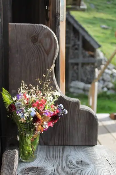 A bouquet of meadow flowers stands on a wooden bench in front of an alpine hut.