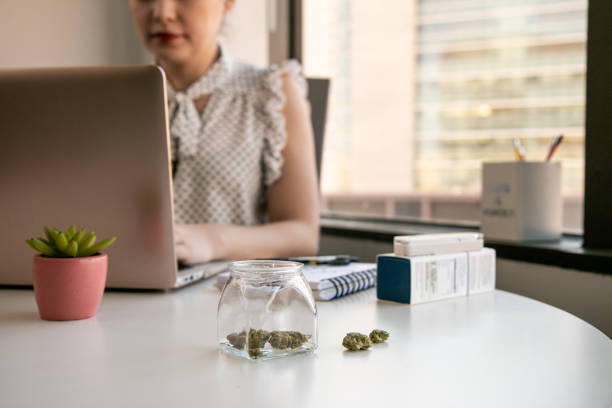 Female Cannabis Entrepreneur working on Marketing for Marijuana Business in Bright, Soft Lit Office Glass jars of cannabis and buds sit on a white office table in an office environment with a downtown view outside the window. A Female Entrepreneur is working on a laptop computer with a vape pen by her side for Marijuana research. A succulent in a pink pot adorns the desk. medical cannabis stock pictures, royalty-free photos & images