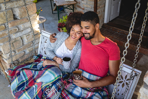 Lovely mixed ethnicity couple enjoying their day under a cozy blanket with some hot tea, on a porch swing.