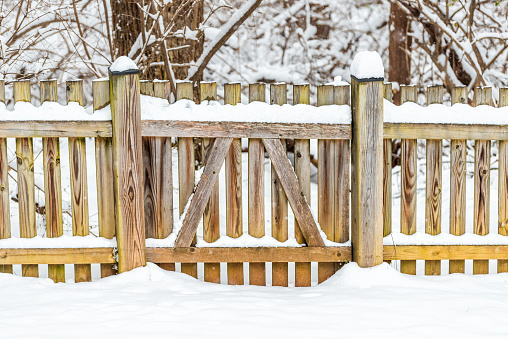 Wooden fence gate locked covered in white snow after heavy snowing snowstorm storm by house home with forest trees bushes in background in Virginia