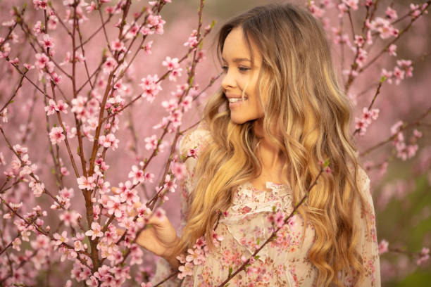 Portrait of amazing and beautiful young woman at spring in orchard of apricot trees in blossom. She is cheerful and happy, enjoying springtime.