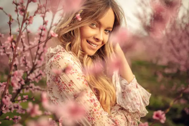 Portrait of amazing and beautiful young woman at spring in orchard of apricot trees in blossom. She is cheerful and happy, enjoying springtime.