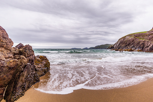 View from small hidden beach towards rocks, sea waves, cliffs and islands in the distance, moody stormy clouds