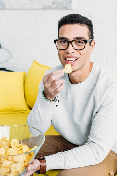 istock handsome young mixed race man sitting on yellow sofa and eating chips 1129487677