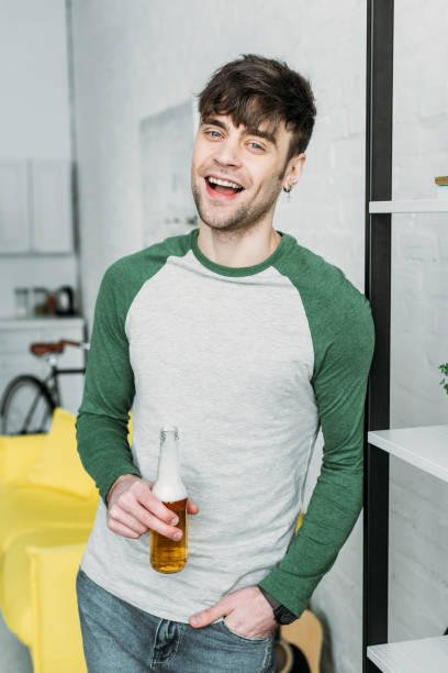 istock smiling handsome man holding bottle of beer while standing by shelving rack 1129487741