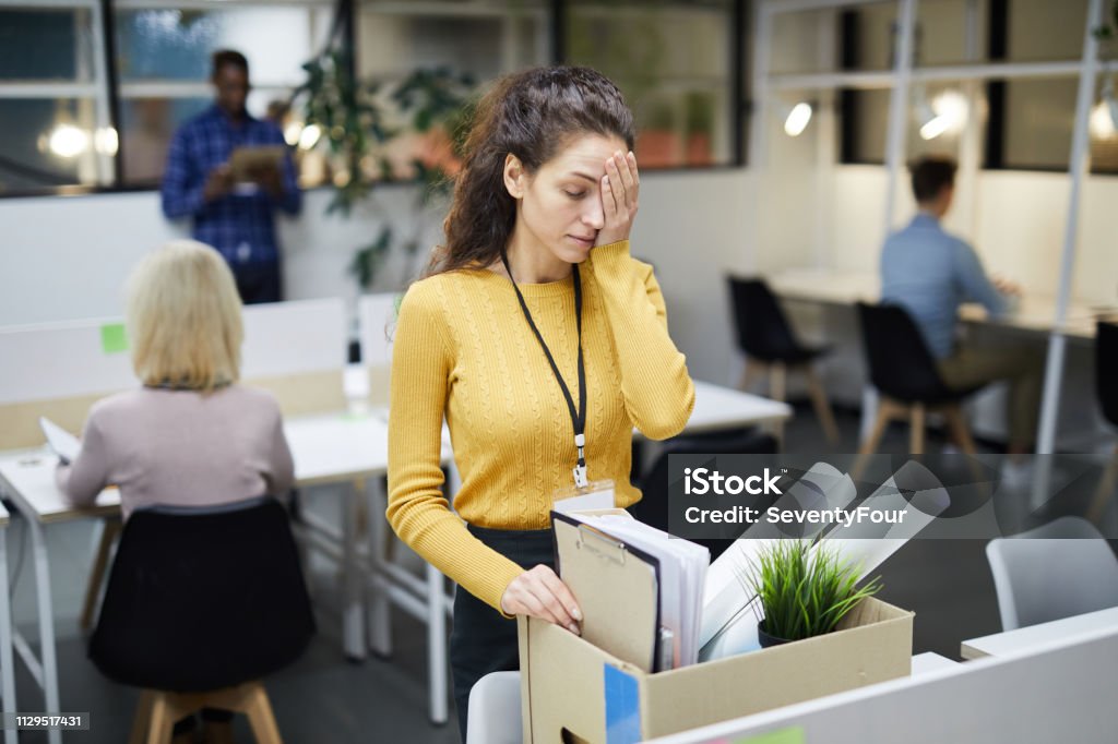 Packing stuff after firing Frustrated young woman in yellow sweater standing at table and touching face with hand while packing stuff in office after dismissal Being Fired Stock Photo