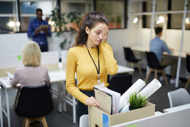 Packing stuff after firing Frustrated young woman in yellow sweater standing at table and touching face with hand while packing stuff in office after dismissal rejection photos stock pictures, royalty-free photos & images