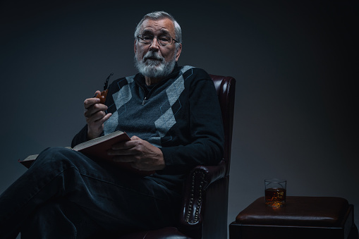 Portrait of a senior male professor sitting in a reading chair with a textbook, pipe and a drink by his side