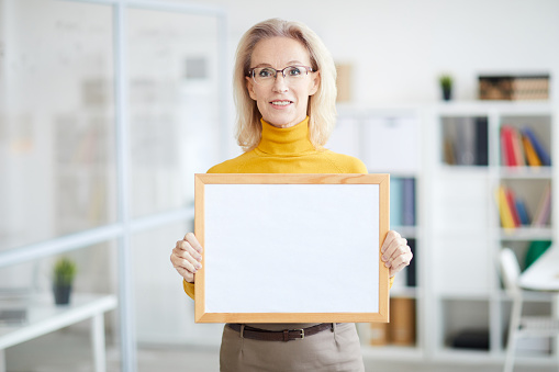 Waist up portrait of smiling mature businesswoman holding blank sign while posing in modern office, copy space