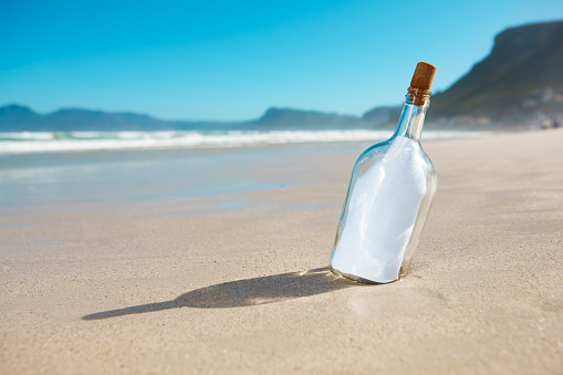 A bottle propped in the sand on a deserted beach contains a blank piece of paper, ready for your message.