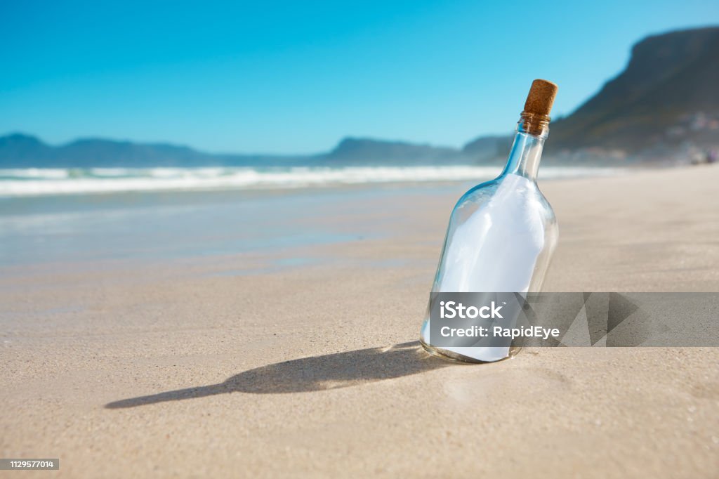 Bottle propped up on sandy beach contains blank note A bottle propped in the sand on a deserted beach contains a blank piece of paper, ready for your message. Bottle Stock Photo
