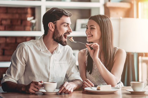 Beautiful couple in love is sitting in cafe, drinking coffee and eating cheesecake. Young woman is feeding her man. Looking softly on each other.