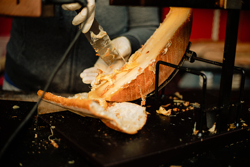 A close-up shot of a raclette cheese wheel in a market, an unrecognizable person can be seen slicing the melting cheese to place onto a slice of bread, the cheese is ready to be served.