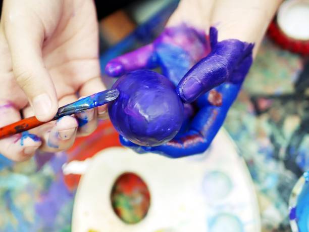 istock Hands of a young student, a brush with paint and a painted egg, a background palette 1129582465