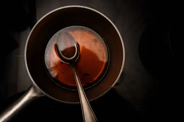 Photo of pot and spoon with beef stock while cooking a red wine morel sauce on a black stove, dark background with copy space, high angle view from above