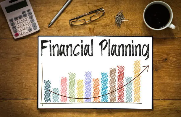 Overhead top view of Financial Planning written in book on desk with a coloured bar chart and trend line.  Desk has coffee cup, glasses, pen and calculator