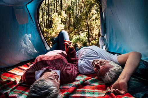 adult senior couple rest lay down inside a tent in free wild camping in the forest for alternative travel and lifestyle. Love forever together concept for man and woman taking hands and look each other - outdoor nature activity