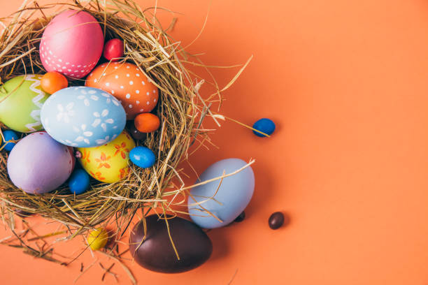 Easter eggs painted by children with chocolate and candies in a hay nest on a orange background. Top view