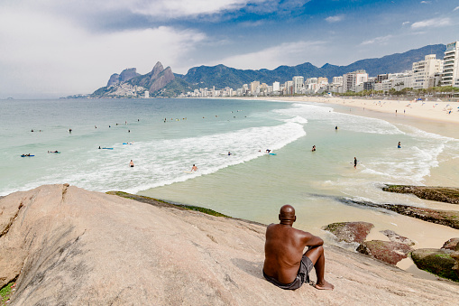 Rio de Janeiro, Brazil - September 14, 2018: Brazilian man sits on rocks at Arpoador Beach watching surfers and enjoying the view of Ipanema