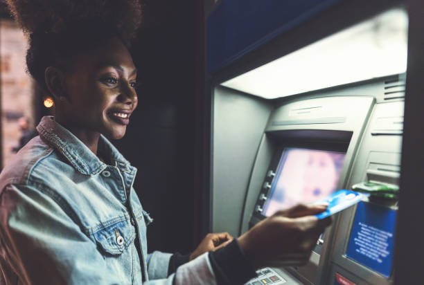 Young woman using ATM device on the street Cheerful African-American woman using her bank card at the ATM in the evening atm photos stock pictures, royalty-free photos & images