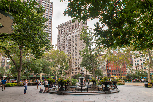 People walking and sitting on benches during summer in Madison Square Park at Midtown Manhattan, New York City, USA. The Flatiron Building at the background.