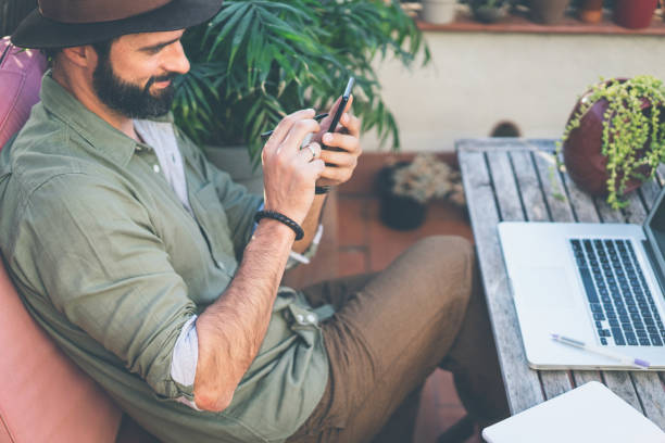 istock Bearded confident hipster wearing green shirt and brown hat texting message via smartphone on terrace outside. Positive man relaxing at summer day. 1129620519