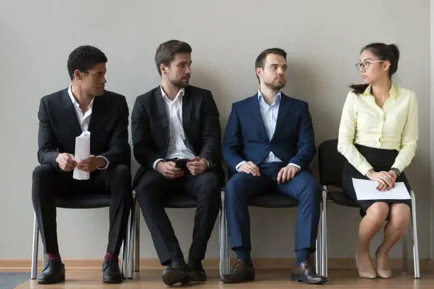 Diverse male applicants looking at female rival among men waiting for at job interview, professional career inequality, employment sexism prejudice, unfair gender discrimination at work concept