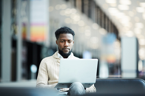 Serious African-american guy concentrating on network in front of laptop while sitting in airport lounge