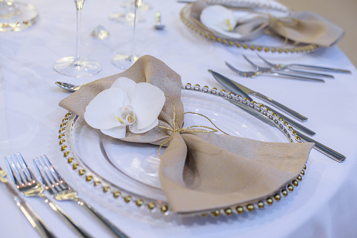 Fine dining table setting featuring transparent plates, beige linen napkin with natural orchid and golden decorations and silverware in the order of use, ready for guests at a formal event or wedding.