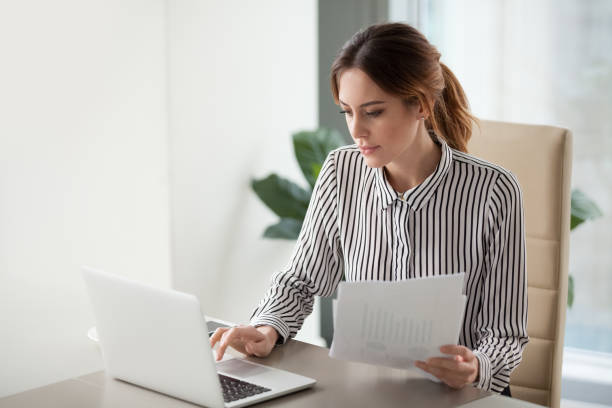 femme d’affaires ciblée grave taper sur ordinateur portable détenant des documents de préparation de rapport - planning manager paperwork business photos et images de collection