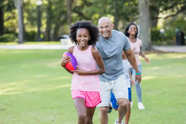 An African-American and Hispanic interracial family spending time together, having fun in the park. The 11 year old girl is holding a football and her father is playfully trying to catch her. Mom and her 9 year old brother are running behind them.