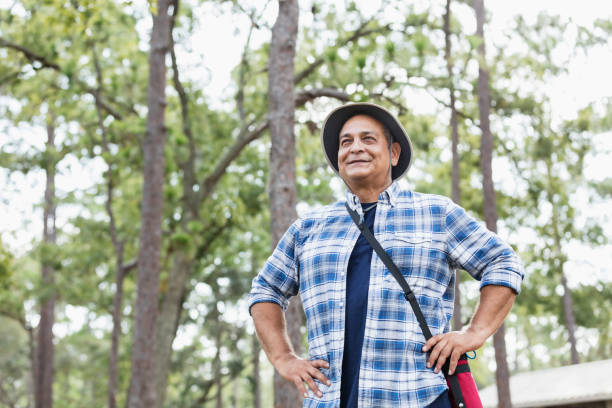 A mature Hispanic man in his 50s enjoying a hike in the park. He is standing, looking at the view of the woodlands, hands on his hips, smiling. He is wearing a plaid shirt and hat, carrying a water bottle.