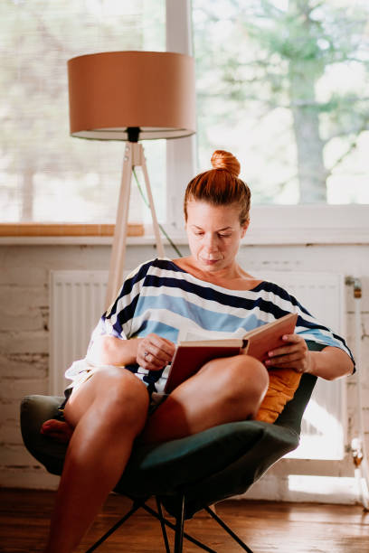 istock Young woman reading a book at home by the window 1129650748