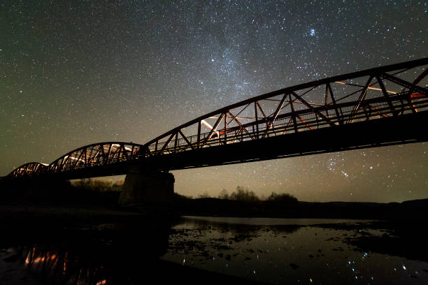 istock Illuminated metal bridge on concrete supports reflected in water on dark starry sky with Milky Way constellation background. Night photography concept. 1129656167