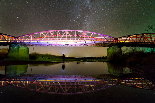 Back view of man with flashlight standing on river bank under illuminated metal bridge under dark starry sky reflected in water. Night photography concept.