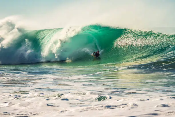 surfer rides by the barrel of a great wave that breaks with a lot of energy and power, sunlight reflects on the turquoise surface of the sea and in the huge pile of foam, wind rises plumes of water