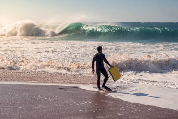 Man on the seashore prepares to surf, wears black neoprene wetsuit, in his hand has a yellow bodyboard. An awesome wave breaks with lots of energy and power, sunlight reflects on the huge pile of foam
