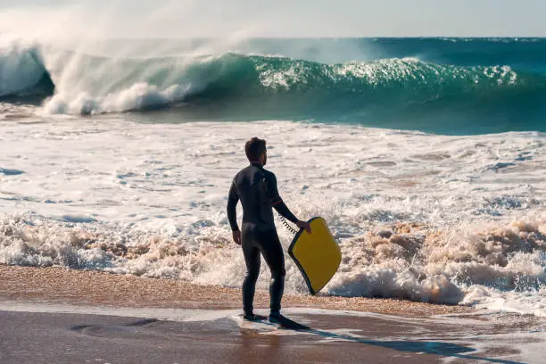 Man on the seashore prepares to surf, wears a black neoprene wetsuit and in his hand has a yellow board. An impressive wave breaks with lots of energy and power, the wind rises plumes of water on it