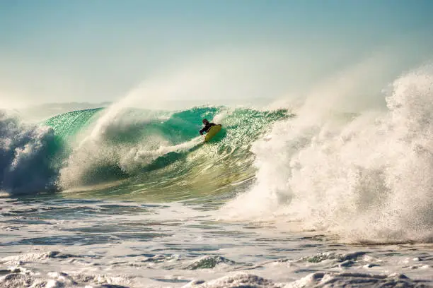 man surfing a big wave that breaks with a lot of energy and power, sunlight reflects golden on the turquoise surface of the sea and in the huge pile of foam, the wind rises plumes of water
