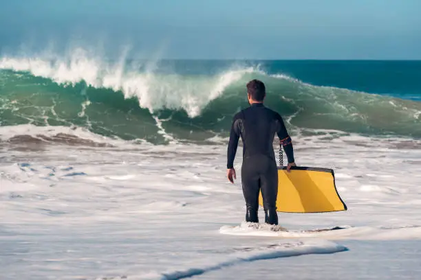 Man on the seashore prepares to surf, wears a black neoprene wetsuit and in his hand wears a yellow bodyboard. the water covers him to the knee while watching a big wave breaking