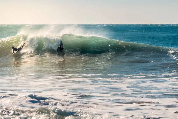 two surfers riding the same beautiful wave, the sunlight reflects gold on foam and the blue and turquoise surface of the sea, the wind raises plumes of water