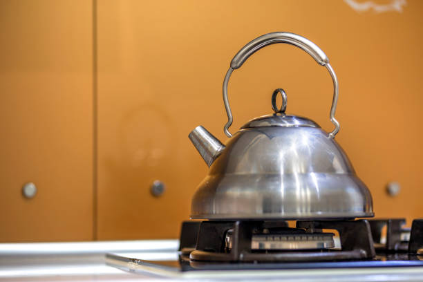 Shiny stainless tea kettle teapot with boiling water on gas stove on kitchen yellow copy space background.