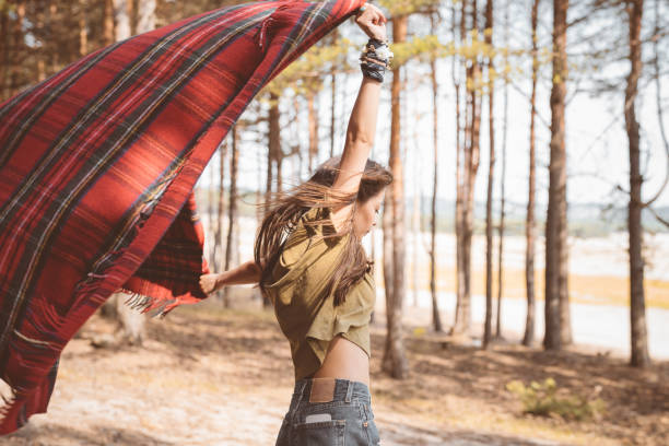 Side view of brunette holding blanket with arms raised. Young female hiker is standing in forest. She is enjoying her summer vacation.