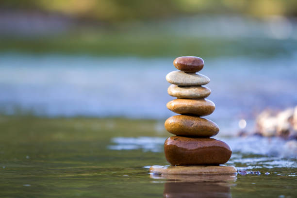istock Close-up abstract image of wet rough natural brown uneven different sizes and forms stones balanced like pyramid pile landmark in shallow water on blurred blue-green misty copy space background. 1129666300