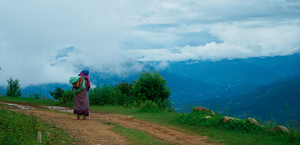 Amppipal, Nepal - August 3, 2017: Single poor nepalese senior woman with backpack and clad in traditional clothing walking barefoot on a dusty country road. Simple life in the nepalese mountains. Near Amppipal village, somewhere in the mountains of the Gorkha district. XXXL (Sony Alpha 7R)