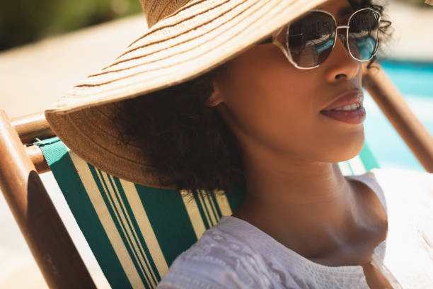 donna afroamericana con cappello e occhiali da sole rilassanti sul lettino nel suo cortile - sun protection foto e immagini stock