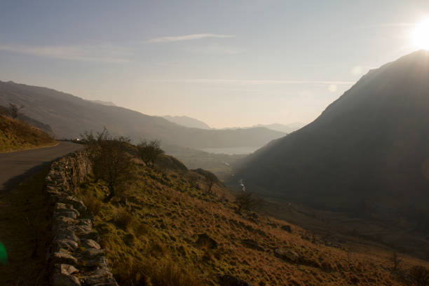 View of road, mountains and lake at sundown. Mountain road, mountain peak, setting sun and a lake in the distance. llyn gwynant stock pictures, royalty-free photos & images