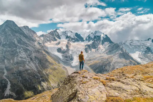 Photo of a bold Young man enjoying the stunning summer view of  peak glacier from St Moritz in Switzerland. Photo of young Traveler Man relaxing meditation with serene view mountains. Landscape Travel Lifestyle hiking concept summer vacations outdoor. Male Day dreamer, human strength and psychology concept.