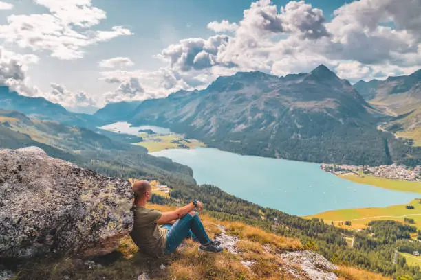 Young man sitting, leaning on big rock while enjoying the stunning summer view of the peak glacier from St Moritz in Switzerland. Photo of  young Traveler Man relaxing meditation with serene view mountains and lake landscape Travel Lifestyle hiking concept summer vacations outdoor. Day dreamer, human strength and psychology concept.